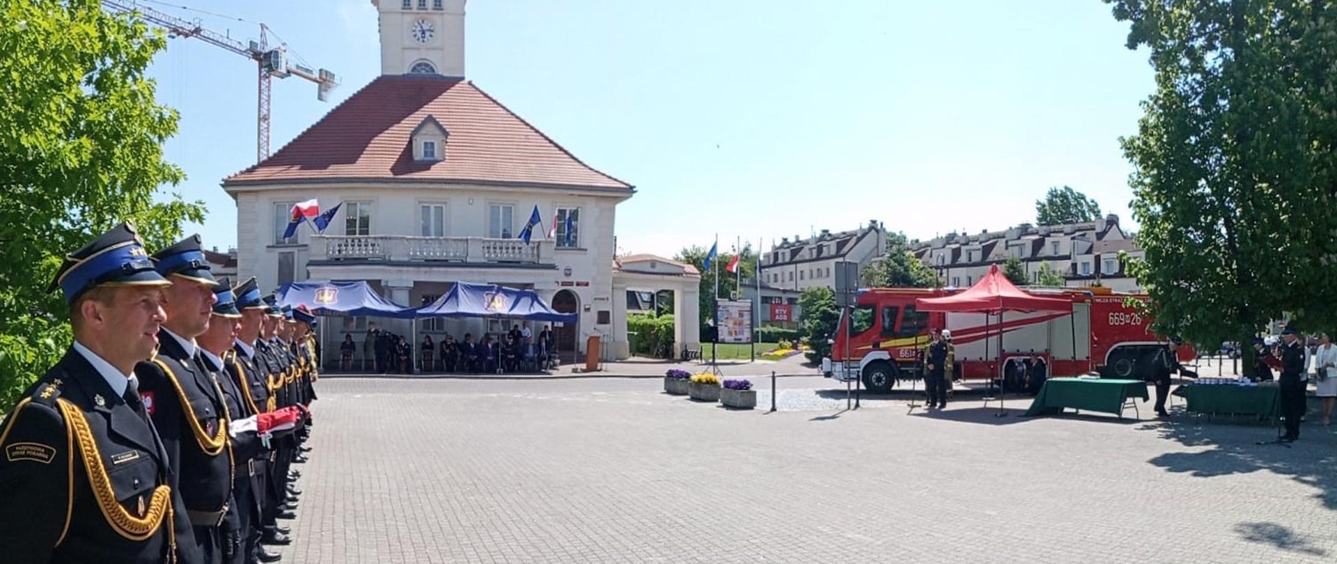funkcjonariusze po lewej stronie ustawieni w rzędzie. w tle widać budynek i przy nim namioty pod którymi stoją goście. po prawej stronie jest samochód ratowniczo-gaśniczy.