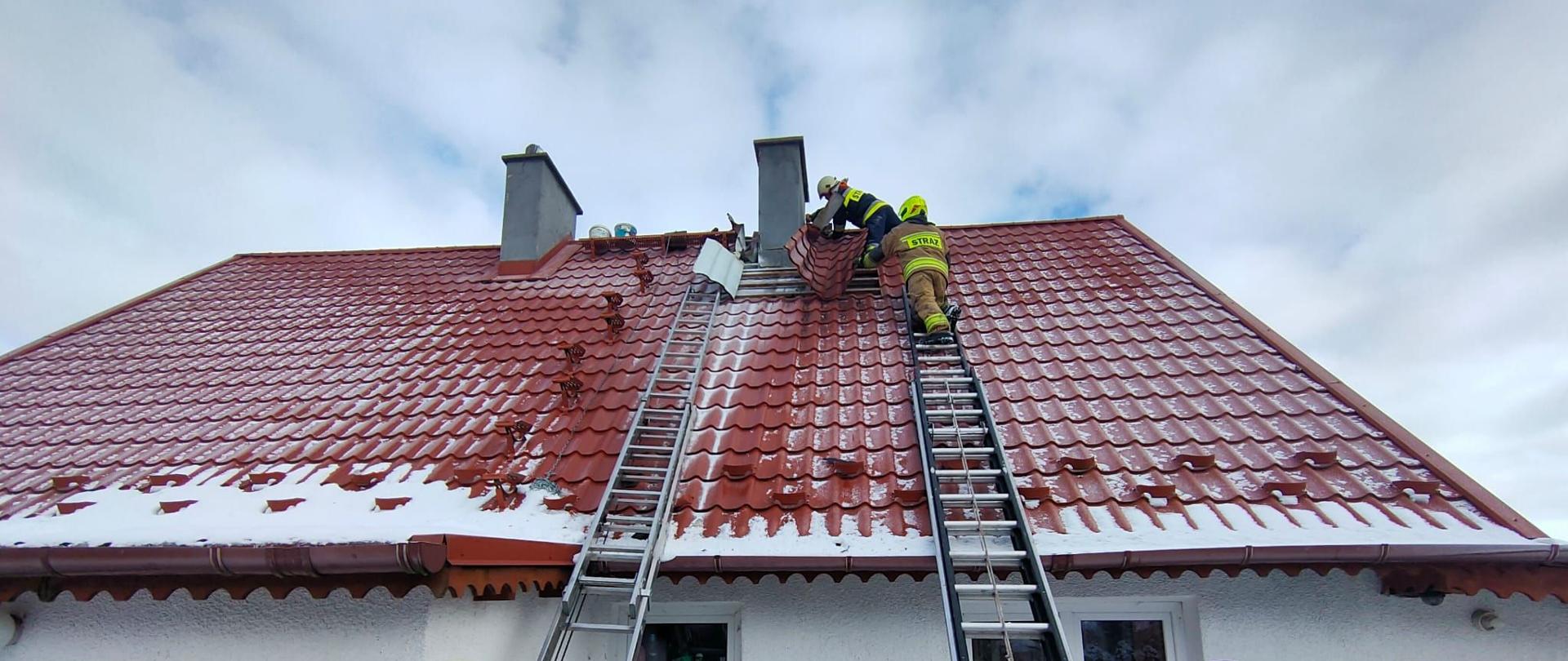 Budynek pokryty czerwona blachodachówką. O dach oparte dwie drabiny strażackie. Na dachu przy kominie siedzi dwóch strażaków. Na dole drabiny trzymają kolejni strażacy.