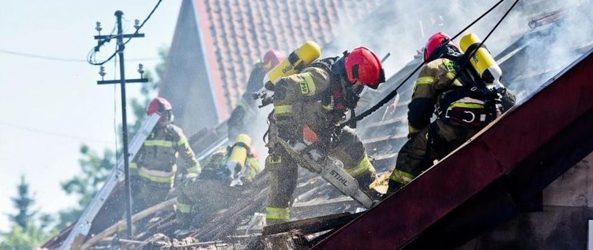 Strażacy podczas prac rozbiórkowych na dachu palącego się wcześniej obiektu. Przy użyciu pilarek mechanicznych i sprzętu burzącego rozbierają spalona konstrukcję dachu.