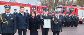 Prezes jednostki OSP Folwarki prezentuje akt włączenia jednostki do KSRG oprawiony w ramce. Na planie zdjęciowym 5 osób. 