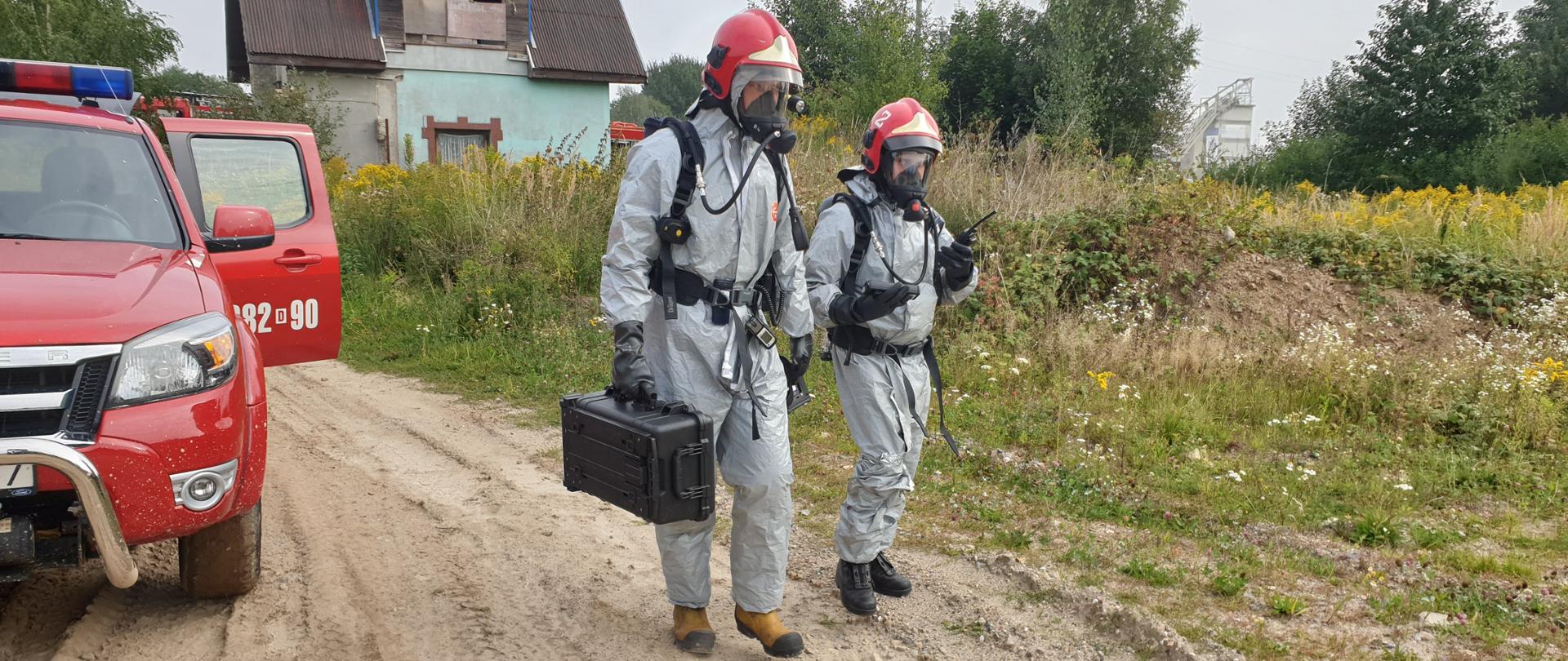 dwóch strażaków ubranych w szare ubrania i aparaty powietrzne. Po lewej stroni stoi samochód strażacki a w tyle widać zielony budynek