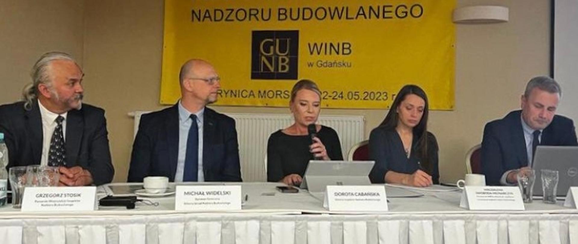 „Pięć osób siedzących przy stole z tabliczkami imiennymi, w tle baner z napisem 'Nadzoru Budowlanego', 'WINB w Gdańsku', datą 22–24.05.2023 i miejscem Krynica Morska.”