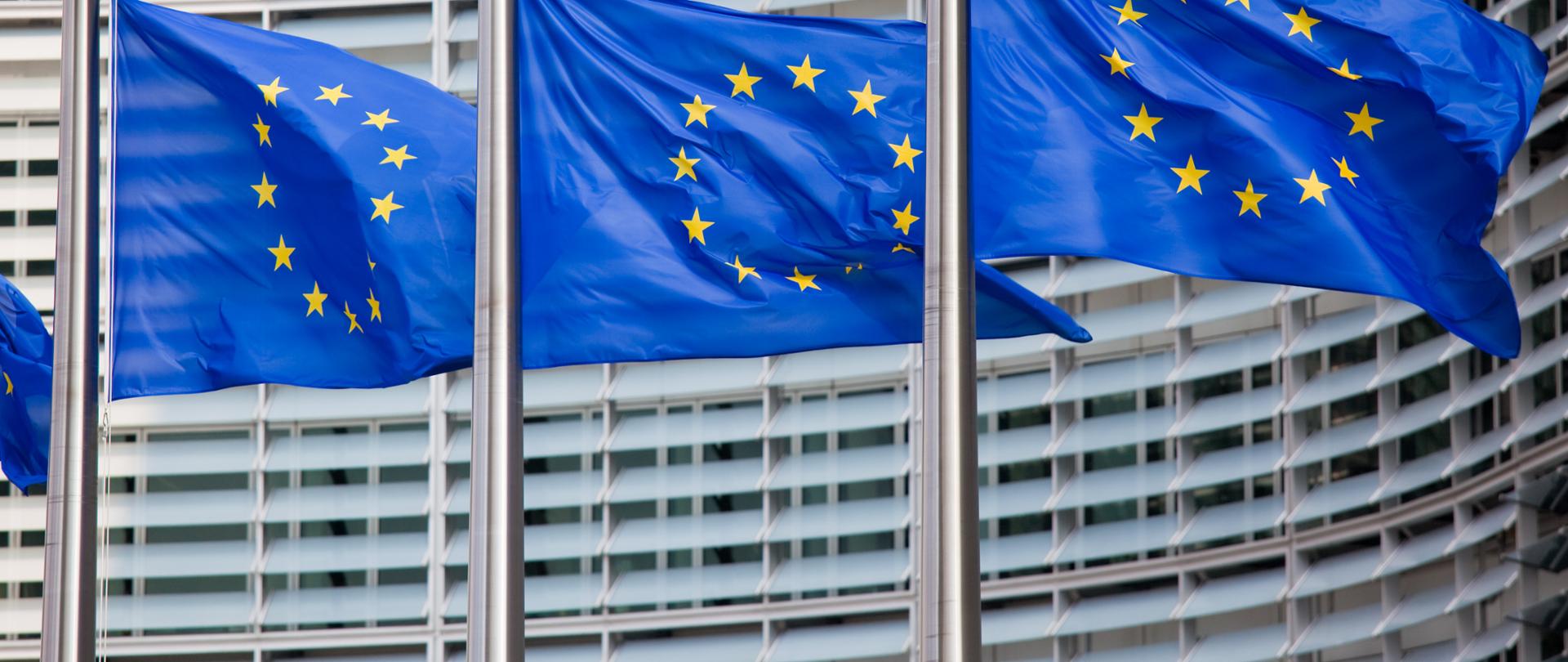 Trzy maszty z flagami Unii Europejskiej stojące na tle gmachu Parlamentu Europejskiego
