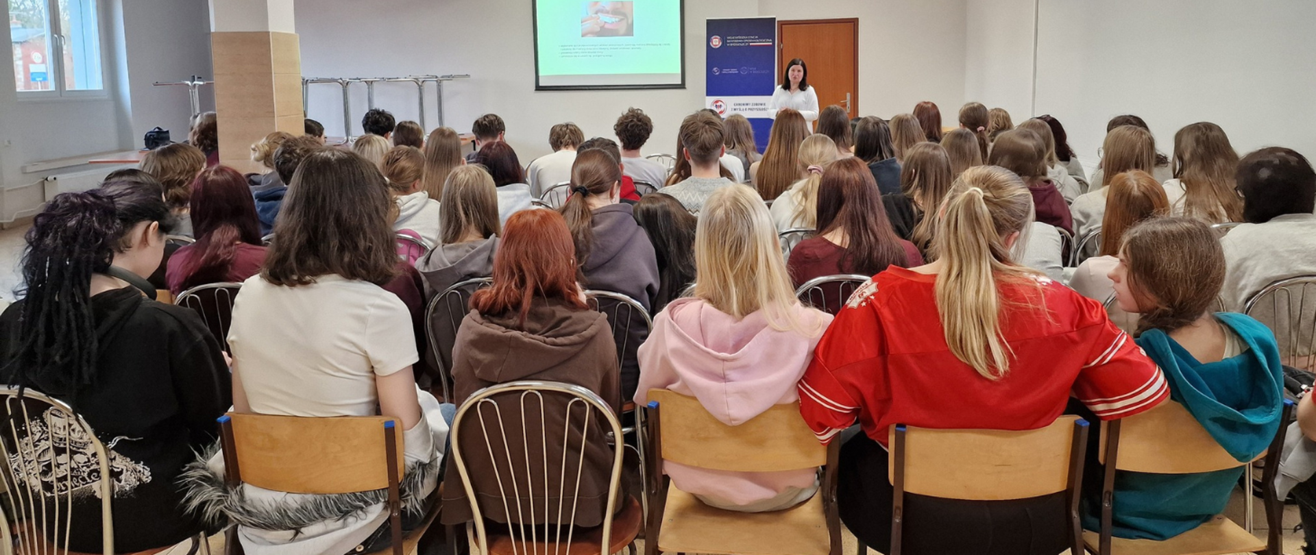 Grupa młodych osób siedzi na krzesłach w sali wykładowej, widziana od tyłu, i słucha kobiety stojącej z przodu przy ekranie z wyświetloną prezentacją.
