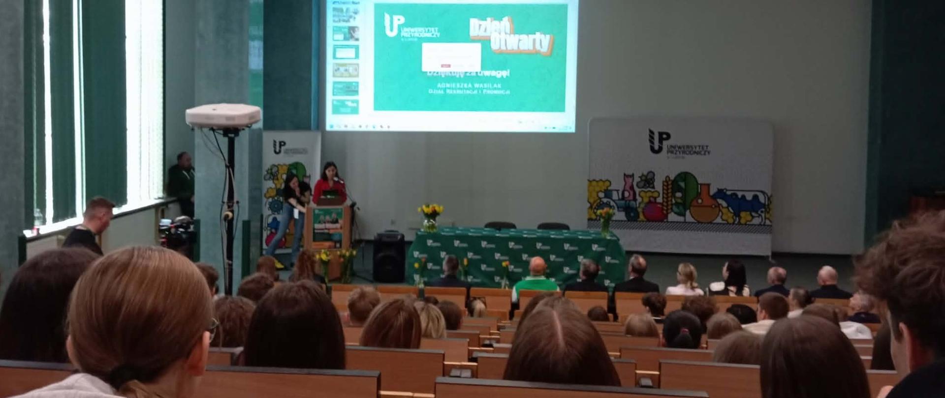 Osoby siedzą w dużej auli i słuchają prezentacji wyświetlanej na ekranie podczas dnia otwartego na uczelni