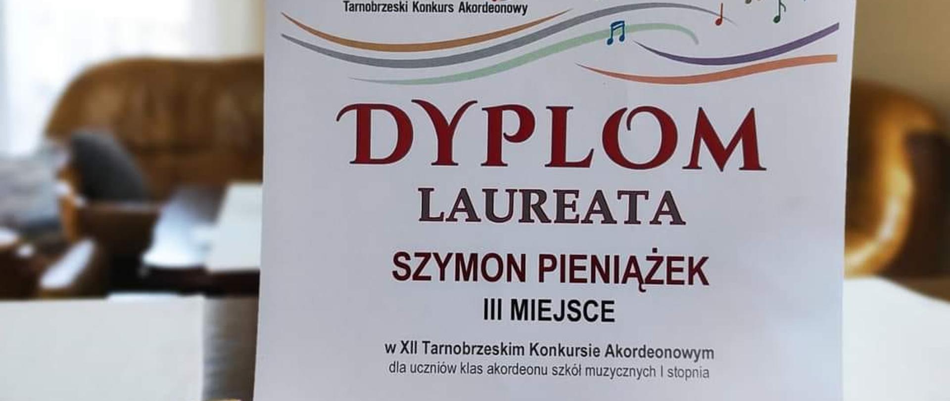 Dyplom Szymona Pieniążka za zajęcie III Miejsca w Tarnobrzeskim Konkursie Akordeonowym, w tle bukiet kwiatów, obok dyplomu statuetka.