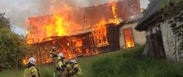 Na zdjęciu płonący dom i strażacy rozwijający linię gaśniczą