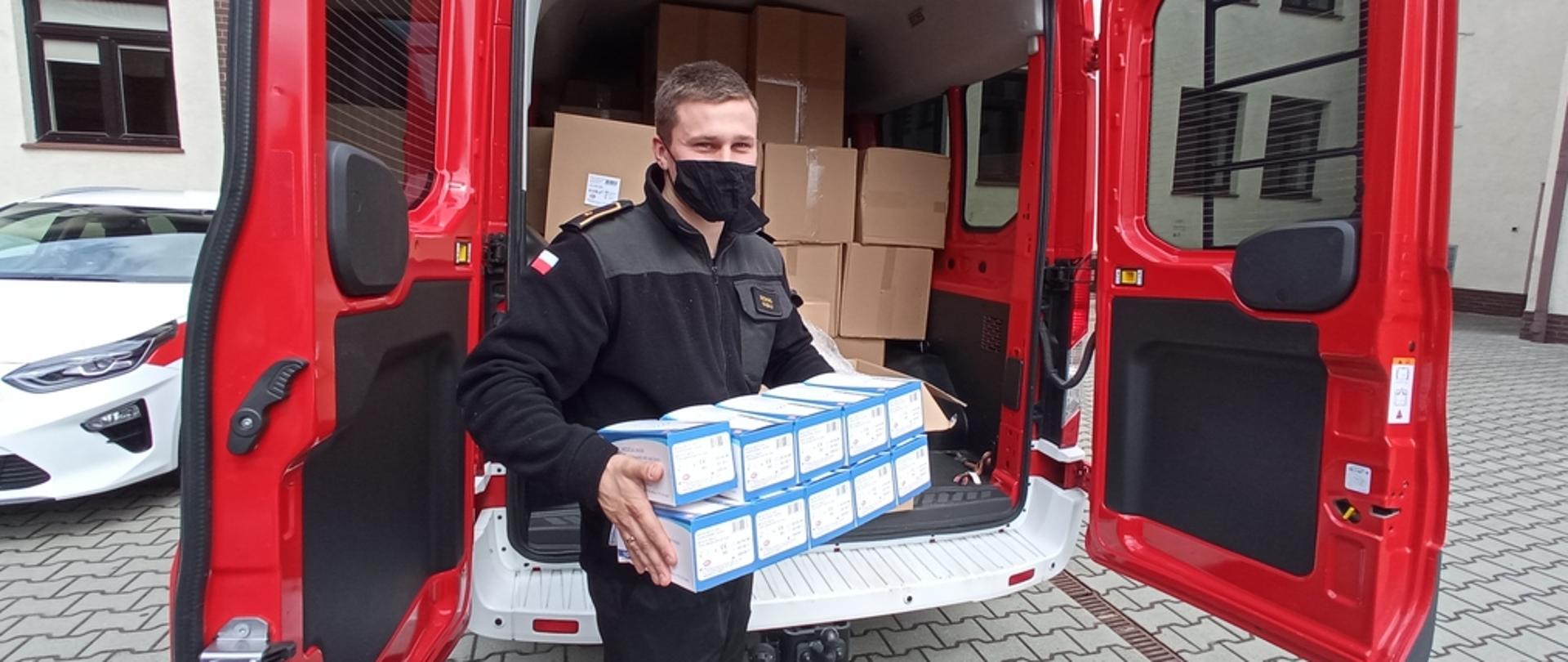 Na zdjęciu strażak w czarnym ubraniu koszarowym podczas wydawania maseczek trzymający w rękach opakowania z maseczkami. Za jego plecami samochód pożarniczy czerwony wypełnimy kartonami z maseczkami ochronnymi.