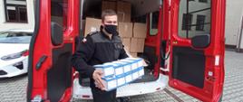 Na zdjęciu strażak w czarnym ubraniu koszarowym podczas wydawania maseczek trzymający w rękach opakowania z maseczkami. Za jego plecami samochód pożarniczy czerwony wypełnimy kartonami z maseczkami ochronnymi.