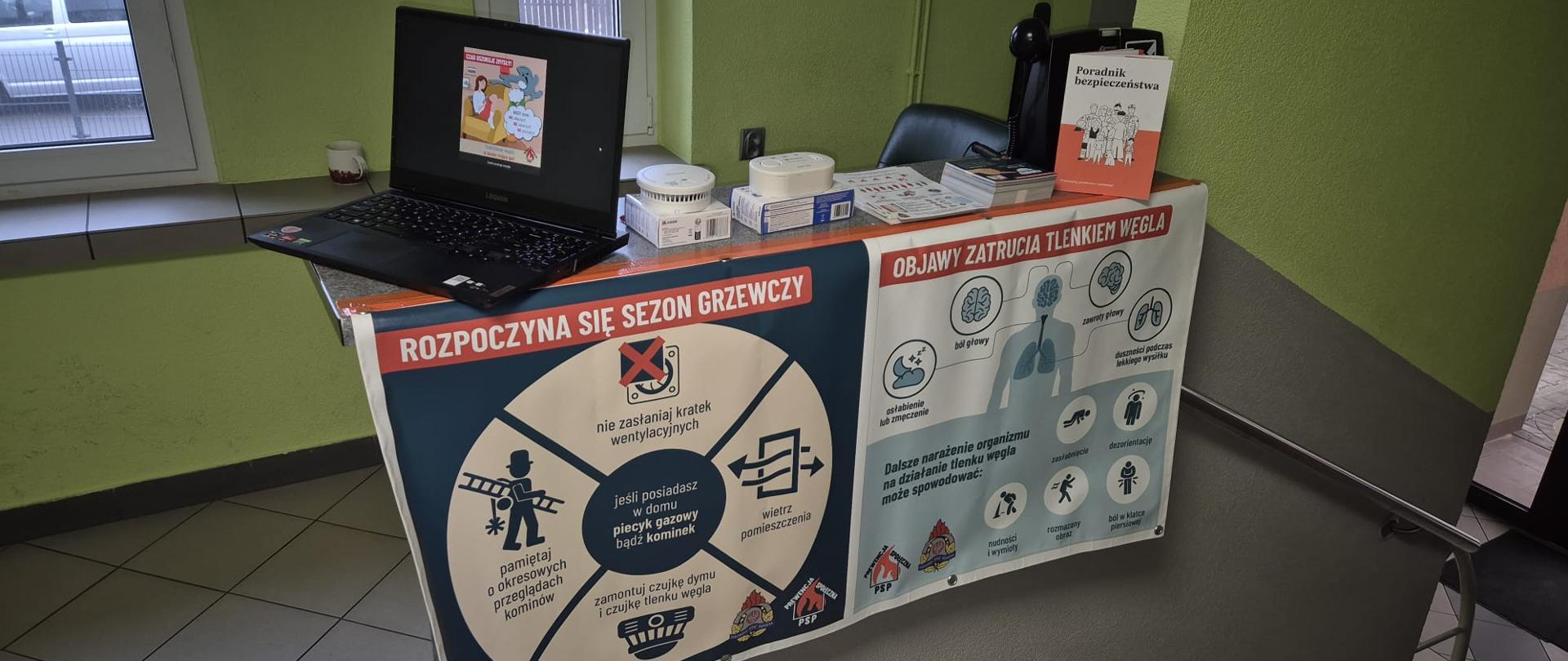Stolik informacyjny ustawiony w korytarzu budynku. Na blacie stoi otwarty laptop z prezentacją, kilka czujników dymu i tlenku węgla oraz broszury o bezpieczeństwie. Przód stolika zasłania duży plakat informacyjny w języku polskim dotyczący sezonu grzewczego i objawów zatrucia tlenkiem węgla, z piktogramami i hasłami ostrzegawczymi. W tle widać zielone ściany, okno oraz schody prowadzące w dół.