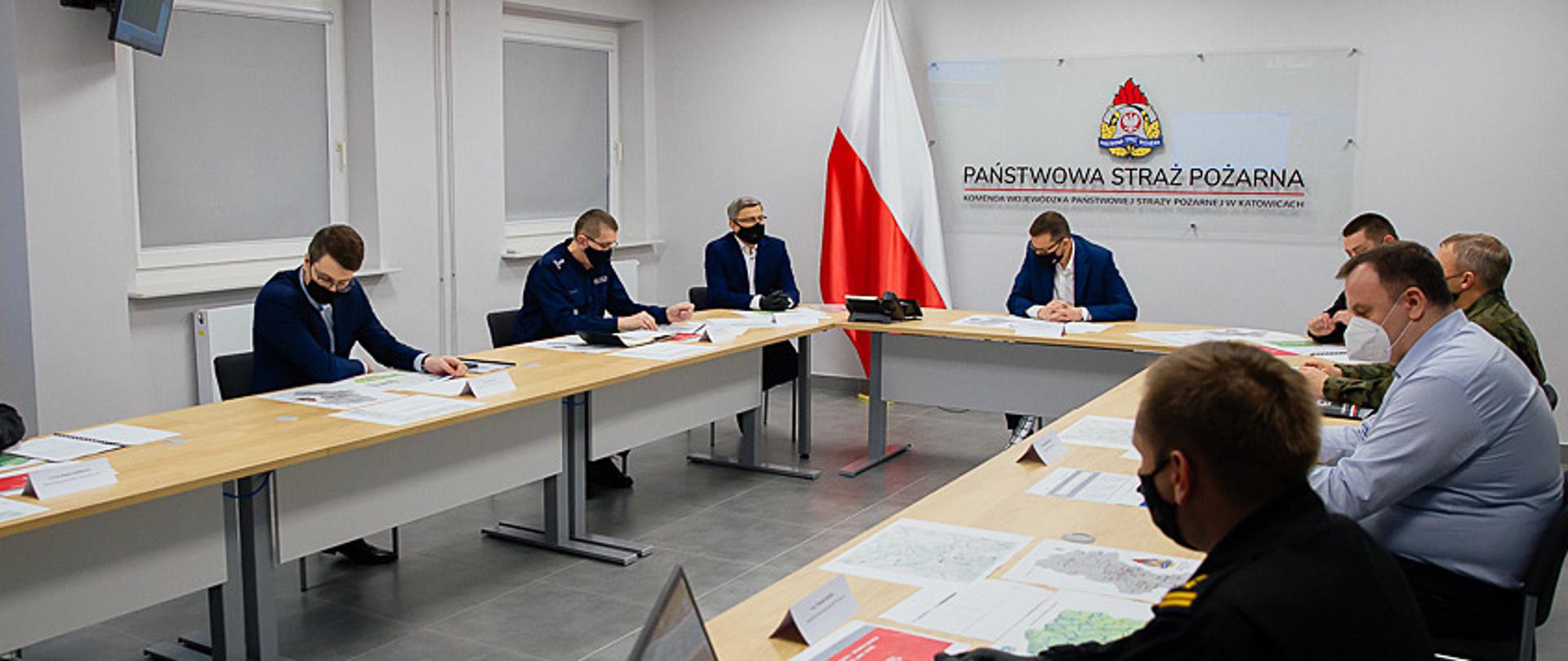 Zdjęcie przedstawia posiedzenie wojewódzkiego zespołu zarządzania kryzysowego w sali sztabowej Komendy Wojewódzkiej PSP w Katowicach. Na zdjęciu widoczny jest siedzący przy stole, w środku premier Matusz Morawiecki, po prawej śląski komendant wojewódzki PSP nadbryg. Jacek Kleszczewski, dowódca 13.Śląskiej Brygady Obrony Terytorialnej płk Tomasz Białas oraz marszałek Województwa Śląskiego Pan Jakub Chełstowski. Po lewej stronie siedzą: wojewoda śląski Jarosław Wieczorek, komendant wojewódzki policji w Katowicach insp. Roman Rabsztyn oraz sekretarz stanu w Kancelarii Prezesa Rady Ministrów Pan Piotr Müller. Uczestnicy posiedzenia przeglądają dokumenty dotyczące sytuacji hydrologicznej. W tle, flaga państwowa, a na ścianie widoczne jest logo PSP , pod nim napis Państwowa Straż Pożarna Komenda Wojewódzka PSP w Katowicach.