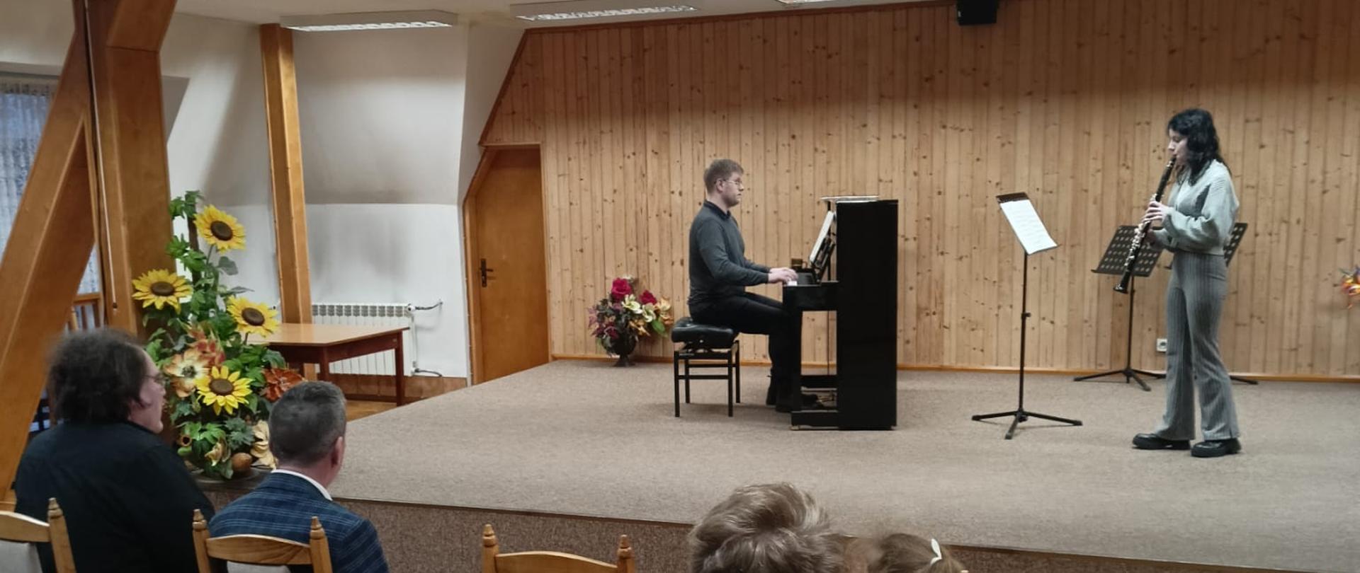 Nastolatka gra na klarnecie, po lewej stronie mężczyzna gra na pianinie. Z przodu fragment widowni z publicznością.