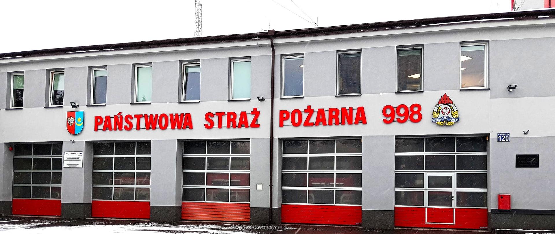 Front budynku jednostki Państwowej Straży Pożarnej z pięcioma bramami garażowymi w czerwono-szarej kolorystyce; nad bramami widoczny napis ‘Państwowa Straż Pożarna 998’ oraz dwa symbole PSP. Plac przed budynkiem pokryty śniegiem.