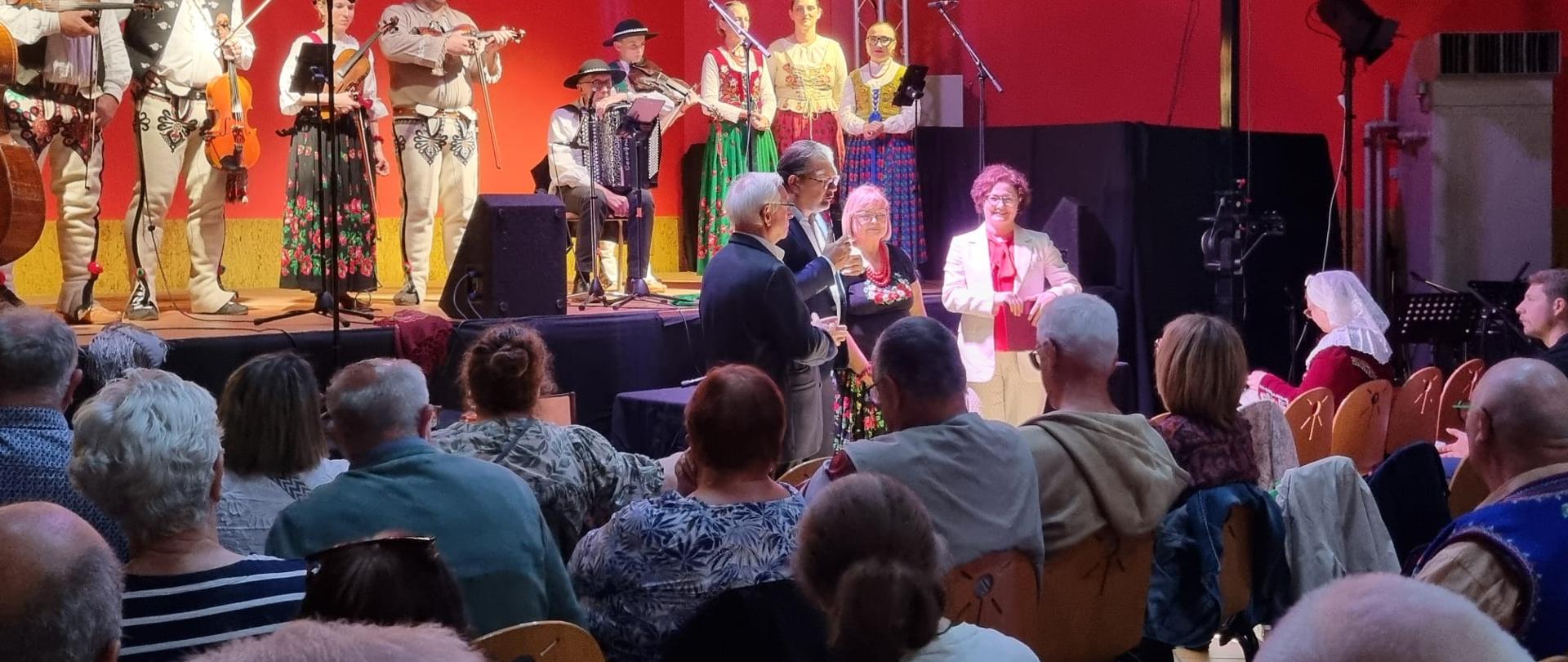 Photo des participants au concert, avec le public assis au premier plan et le groupe de musiciens en costumes folkloriques polonais visible à l'arrière-plan.