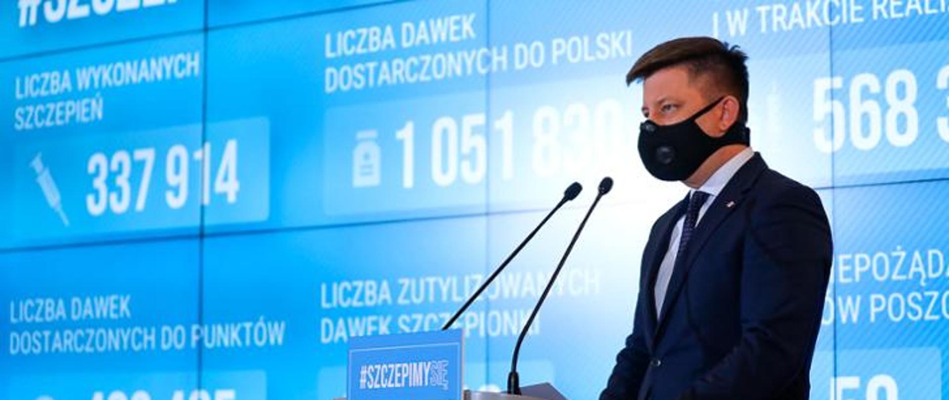 Minister Michał Dworczyk podczas konferencji prasowej. Przemawia do mikrofonów. W tle ekran na którym wyświetlane są statystyki dotyczące wykonanych szczepień przeciw Covid-19.