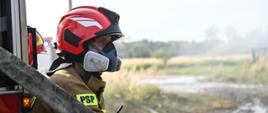 Strażak w czerwonym hełmie z białymi odblaskami, w masce na twarzy. W ubraniu specjalnym, na ramieniu napis PSP. Cześciowo zasłonięty przez odcinek węża tłocznego. W tle rozlewisko piany gaśniczej.