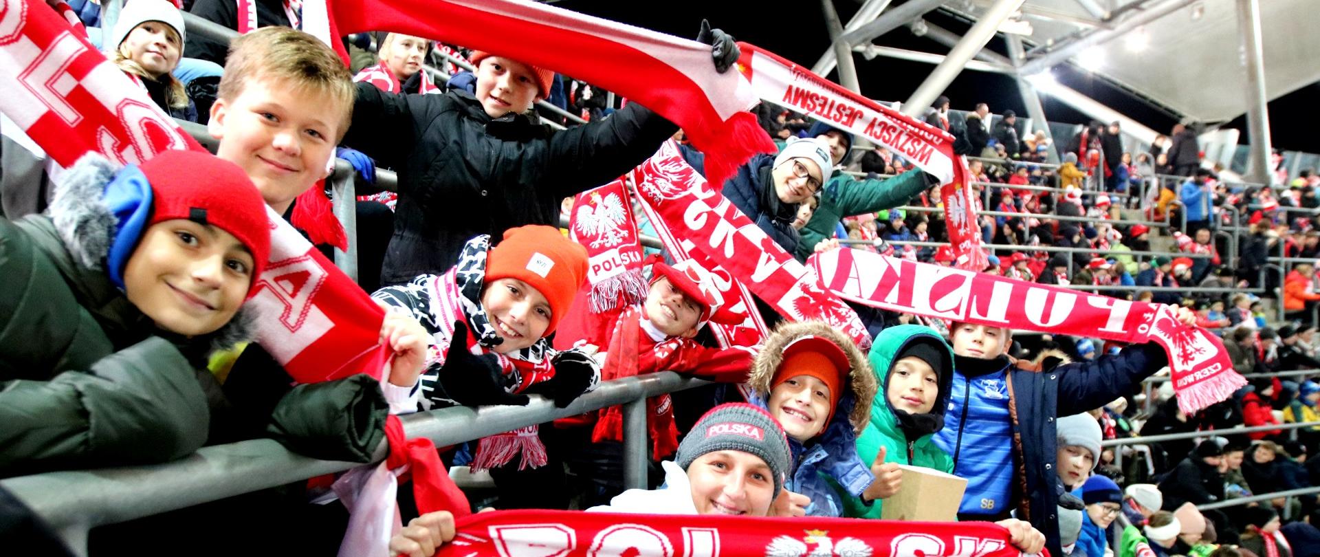 Duża grupa roześmianych dzieci siedzi na trybunie stadionu, machają biało-czerwonymi szalikami.