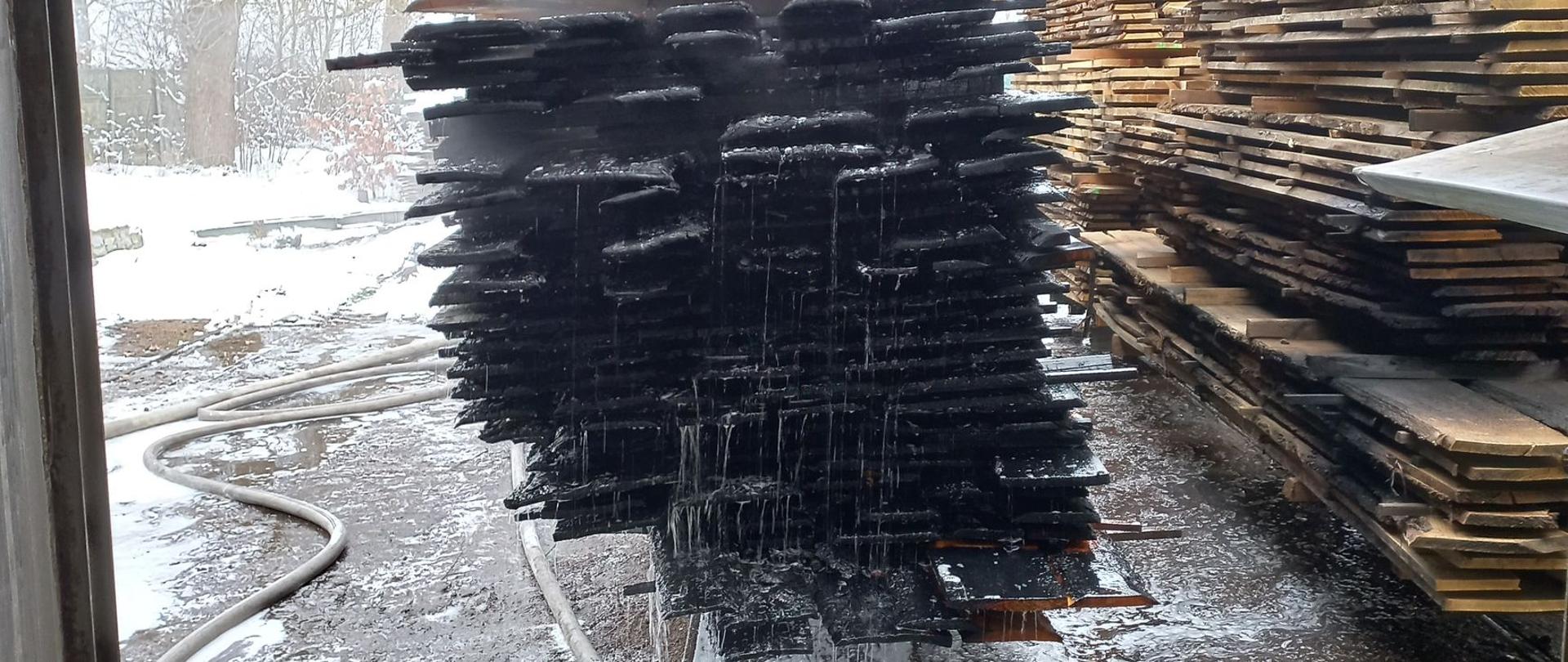 Zdjęcie przedstawia spalone deski na terenie tartaku, gdzie doszło do pożaru.
W tle inne deski i drzewa.
