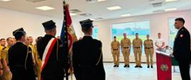 W nowoczesnej, jasnej sali odbywa się uroczysty apel służbowy. Na pierwszym planie stoi poczet sztandarowy w granatowych mundurach galowych – jeden z funkcjonariuszy trzyma sztandar z czerwono‑białymi barwami i złotymi elementami. Obok niego pozostali członkowie pocztu stoją w równym szyku. W głębi sali widoczna jest grupa strażaków w piaskowych mundurach, ustawionych w szeregu na baczność. Po prawej stronie kadru znajduje się mównica z emblematem straży pożarnej przedstawiającym stylizowany płomień i skrzyżowane topory; za mównicą stoi funkcjonariusz w ciemnym mundurze, prowadzący uroczystość. Z boku obserwują ją również osoby cywilne, w tym dziecko. Całość tworzy podniosłą, oficjalną atmosferę podkreślającą rangę wydarzenia.