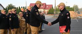 Strażacy stoją ustawieni w dwóch rzędach. Przed nimi na środku stoi dwóch strażaków oddają sobie honor przez podanie ręki. W tle zabudowa osiedlowa i plac budowy.