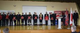 Wspólne zdjęcie strażaków zaproszonych gości oraz przedstawicieli Młodzieżowych Drużyn Pożarniczych którzy trzymają czerwone teczki.