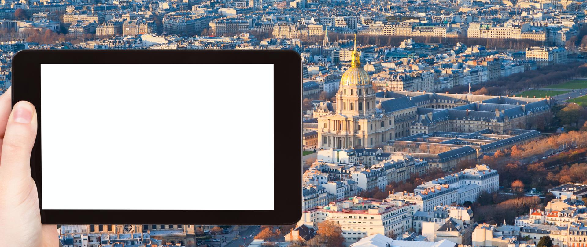 Tablet z białym ekranem trzymany w lewej dłoni na tle panoramy miasta