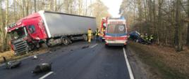 Zdjęcie przedstawia samochód ciężarowy z uszkodzonym przodem znajdujący się częściowo w rowie oraz samochód osobowy marki Opel znajdujący się w przydrożnym rowie. Samochód osobowy z uszkodzonym przodem oraz wyciętym dachem przez strażaków w celu ewakuacji poszkodowanej kobiety. Na zdjęciu widać samochód strażacki , karetkę pogotowia ratunkowego oraz strażaków i policjantów.