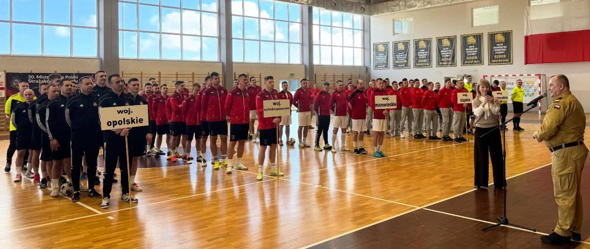 Przemówienie rozpoczynające turniej. Na hali sportowej stoją reprezentacje 5 województw. 