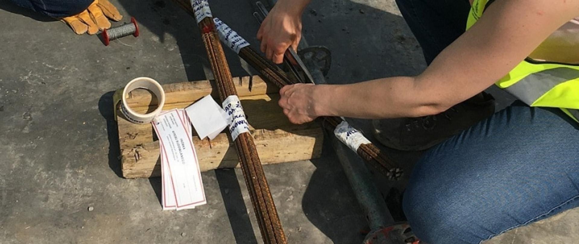 Osoba w kamizelce odblaskowej pracująca przy zbrojeniu betonowej powierzchni z użyciem prętów i taśmy.