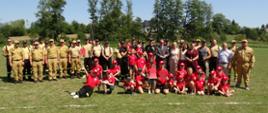 Zdjęcie grupowe uczestników zawodów pożarniczych – strażacy w mundurach i strojach sportowych oraz dziecięce i młodzieżowe drużyny w czerwonych koszulkach pozują na boisku z pucharami i dyplomami.