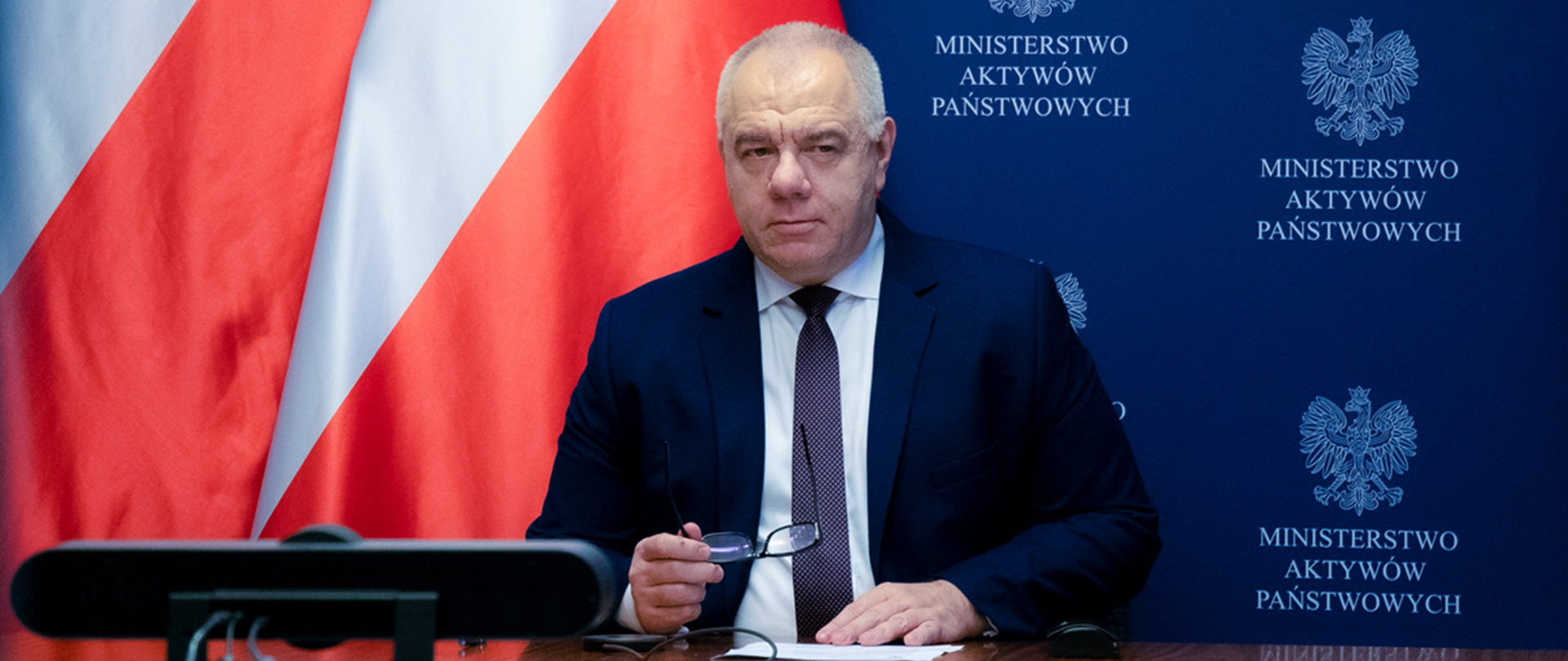 Wicepremier Jacek Sasin podczas spotkania online. Siedzi przy biurku, przed nim stoi kamera internetowa. Minister trzyma w dłoni okulary. W tle ścianka MAP.