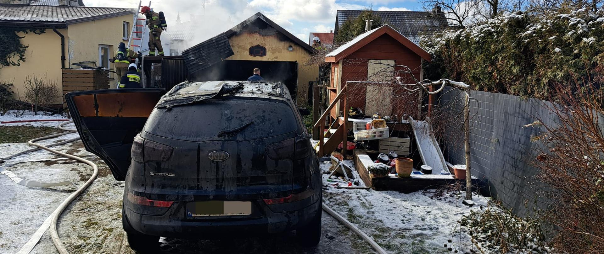 Zdjęcie przedstawia samochód osobowy, który uległ spaleniu stojący przed garażem gdzie doszło do pożaru. Po lewej stronie widać dom mieszkalny, a po prawej obiekt małej architektury.