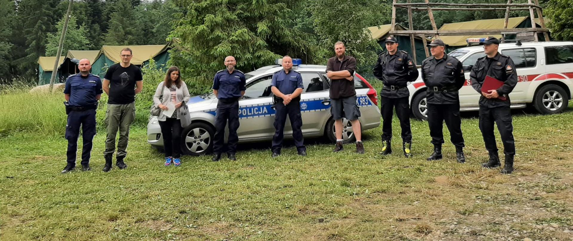 Na zdjęciu widać funkcjonariuszy Państwowej Straży Pożarnej, Policji oraz opiekunów harcerzy którzy stoją przed samochodami straży i policji. W oddali widać namioty harcerskie oraz las.