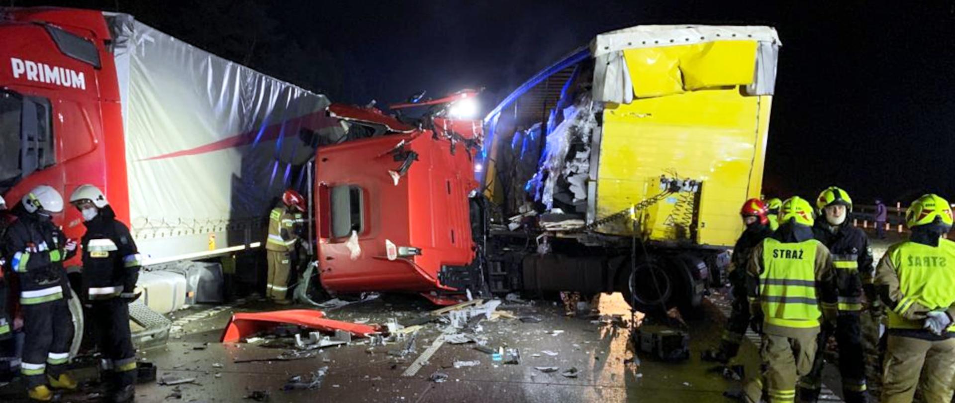 Zdjęcie przedstawia dwa samochody ciężarowe po zderzeniu. Wokół widać grupy strażaków wykonujące czynności ratownicze.