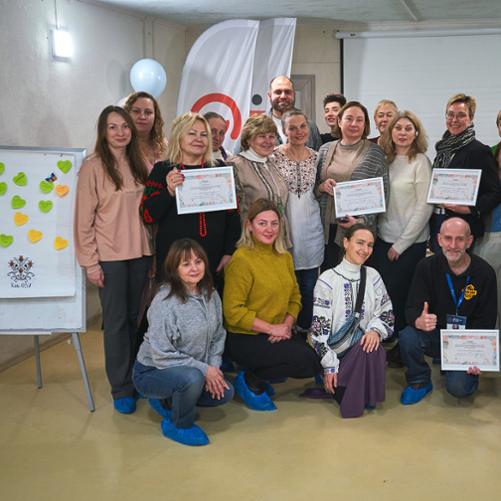 Grupa kobiet, część w ukraińskich strojach ludowych, prezentuje certyfikaty, w tle widać zarys logo Polskiej pomocy