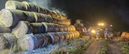 Nocne zdjęcie plenerowe ukazuje duży stos cylindrycznych bel słomy, z których niektóre wydają się płonąć lub tlić, wydzielając dym unoszący się w ciemne niebo. Na prawo od płonącego siana widać kilku strażaków w odzieży ochronnej, oświetlonych jasnymi światłami pojazdu, ciągnika z turem, zaparkowanego w pobliżu. Wyglądają na aktywnie zaangażowanych w akcję gaśniczą, z widocznymi wężami na ziemi. Przed belami słomy, w kierunku strażaków, biegnie polna ścieżka. Cała scena przedstawia pożar produktów rolnych.