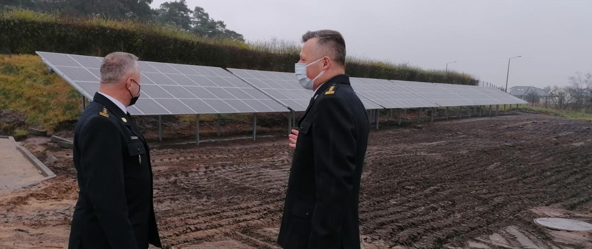 Dwaj strażacy w mundurach galowych i założonych maseczkach stoją bokiem do widza a twarzami do siebie na utwardzonym placu i rozmawiają. W tle za nimi znajdują się: wyrównana ziemia oraz długi rząd paneli fotowoltaicznych posadowionych na metalowej konstrukcji przy kilkumetrowej skarpie na szczycie której rośnie zielony żywopłot. 