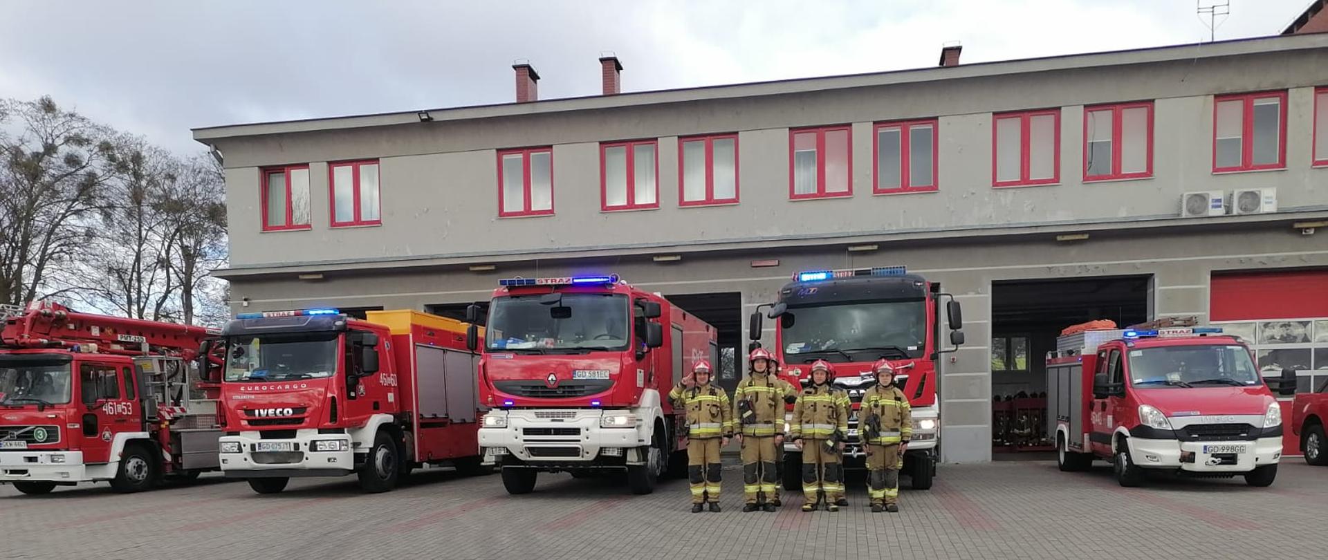 Samochody bojowe z włączoną sygnalizacją alarmową oraz strażacy stoją przed budynkiem komendy