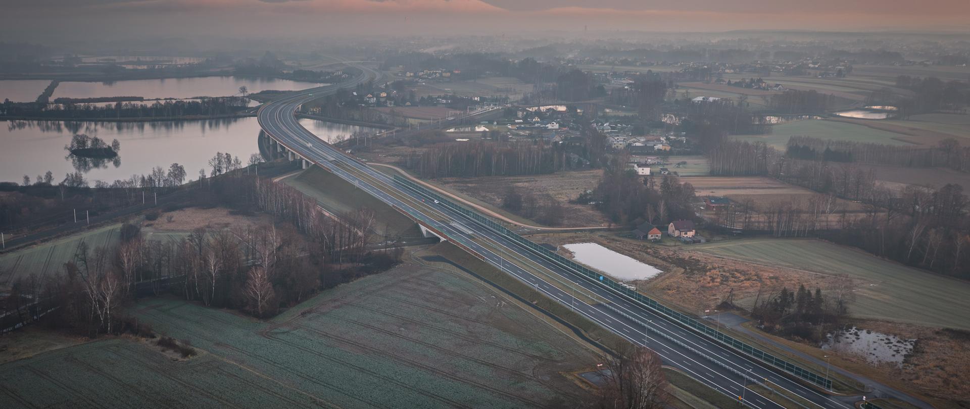 Zdjęcie przedstawia widok z lotu ptaka na drogę ekspresową biegnącą przez wiejski krajobraz o świcie lub zmierzchu. Po lewej stronie widoczne są rozległe zbiorniki wodne, a w tle rozciąga się panorama miejscowości z rozproszoną zabudową.