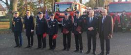 Zdjęcie przedstawia zasłużonych strażaków OSP, którym przyznano świadczenie ratownicze w towarzystwie Komendanta Wojewódzkiego PSP, Wojewody Pomorskiego oraz posłów na Sejm RP, którzy pozują na tle samochodów strażackich.