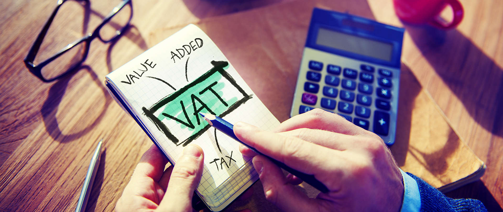 Notes na którym widnieje napis VAT w tle widać blat stołu na którym jest kalkulator okulary i długopis