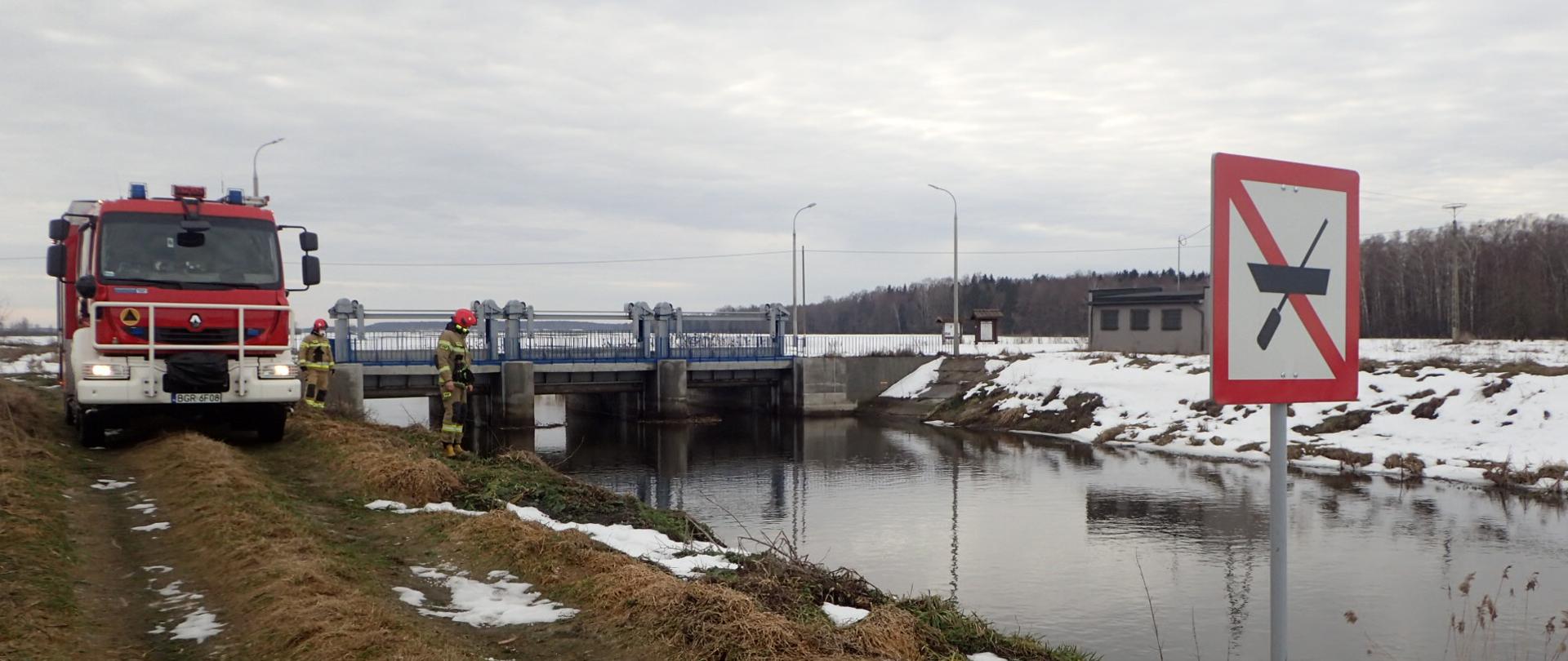 Na zdjęciu strażacy PSP sprawdzają stan wody w rzece. w tle samochód gaśniczy PSP.