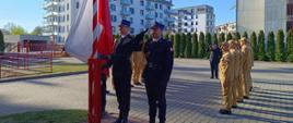 Trzech strażaków w ciemnych mundurach wyjściowych i czapkach rogatywkach stoi przy biało-czerwonym maszcie. Jeden z nich salutuje, drugi trzyma flagę. W tle nowoczesna zabudowa mieszkaniowa i stojący w szeregu strażacy w piaskowych mundurach służbowych.