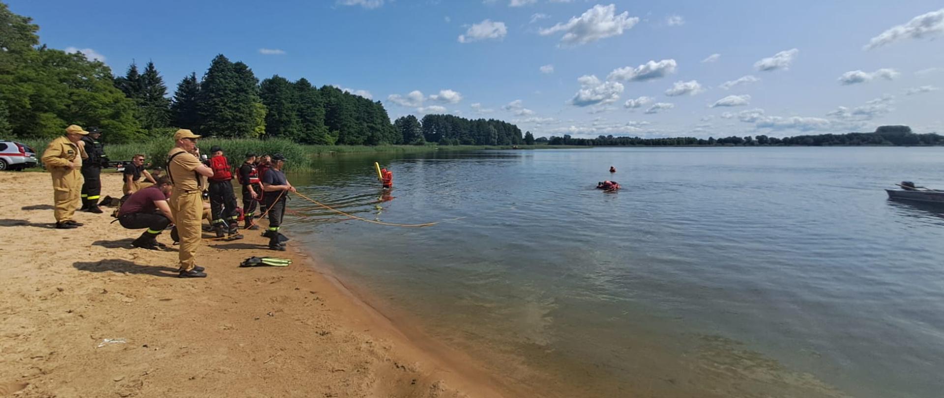 Ćwiczenia z zakresu ratownictwa wodnego. Zdjęcie przedstawia strażaków PSP i OSP ubranych w suche skafandry do ratownictwa wodnego podczas ćwiczeń na akwenie Jeziora Głuszyńskiego