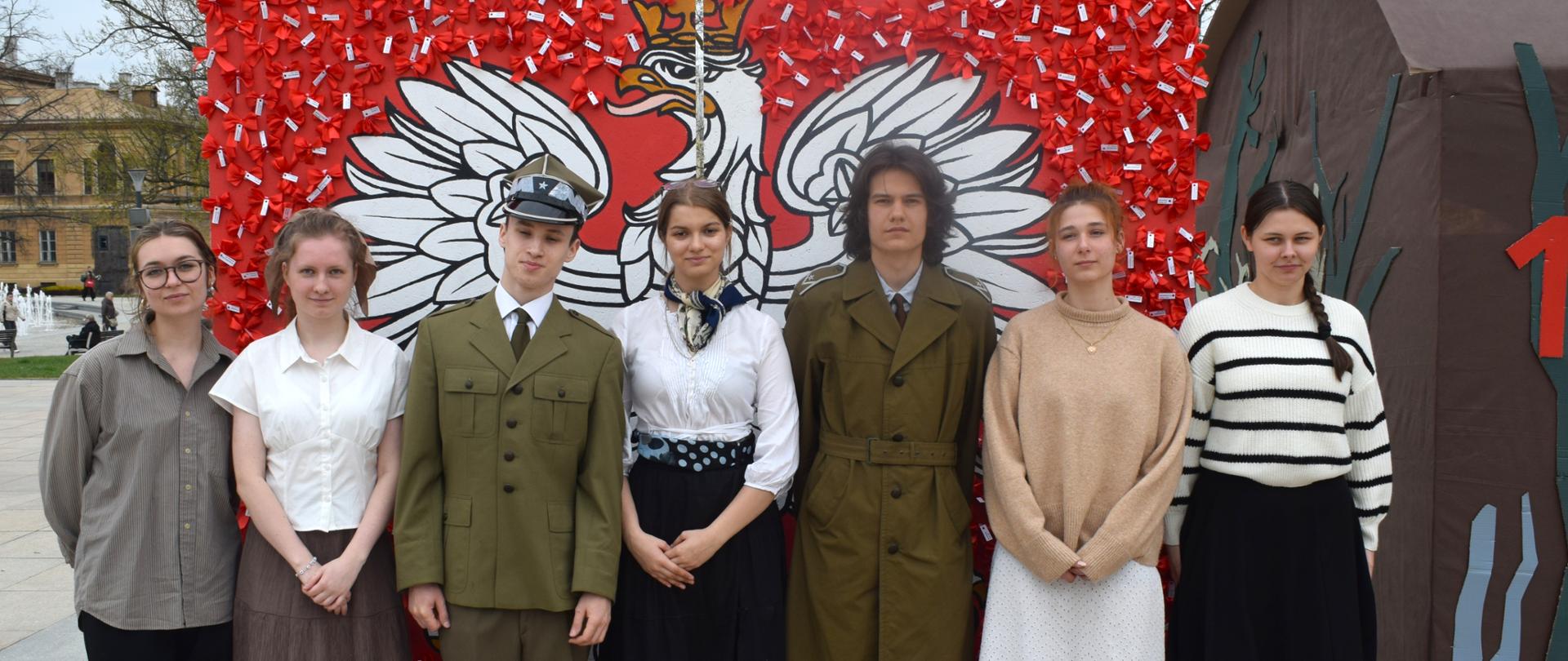 Duże godło Polski (czerwone tło i biały orzeł) na placu miejskim (płyty chodnikowe, z tyłu drzewa). Przed godłem, pozując do zdjęcia, młode osoby, dwie z nich w mundurach