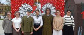 Duże godło Polski (czerwone tło i biały orzeł) na placu miejskim (płyty chodnikowe, z tyłu drzewa). Przed godłem, pozując do zdjęcia, młode osoby, dwie z nich w mundurach