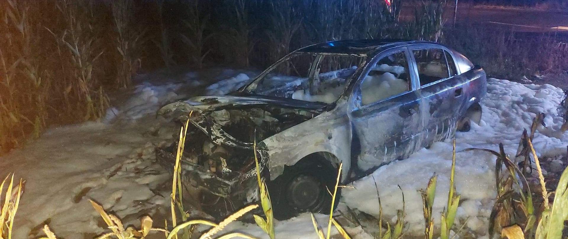 Zdjęcie przedstawia spaliny całkowicie samochód osobowy marki Opel. Pora nocna. Samochód zaparkowany na polu kukurydzy. Wewnątrz samochodu oraz wokół niego widać pianę gaśniczą. 