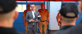 Mężczyzna w garniturze stoi przed mikrofonem za nim stoją strażacy Państwowej Straży Pożarnej mężczyzna przemawia do strażaków jednostki ratowniczo-gaśniczej.