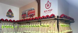 Pomieszczenie ze strojami strażackimi dla dzieci. Na ścianie logo Komendy Powiatowej PSP w Pleszewie, logo Państwowej Straży Pożarnej oraz rysunek pojazdu pożarniczego.