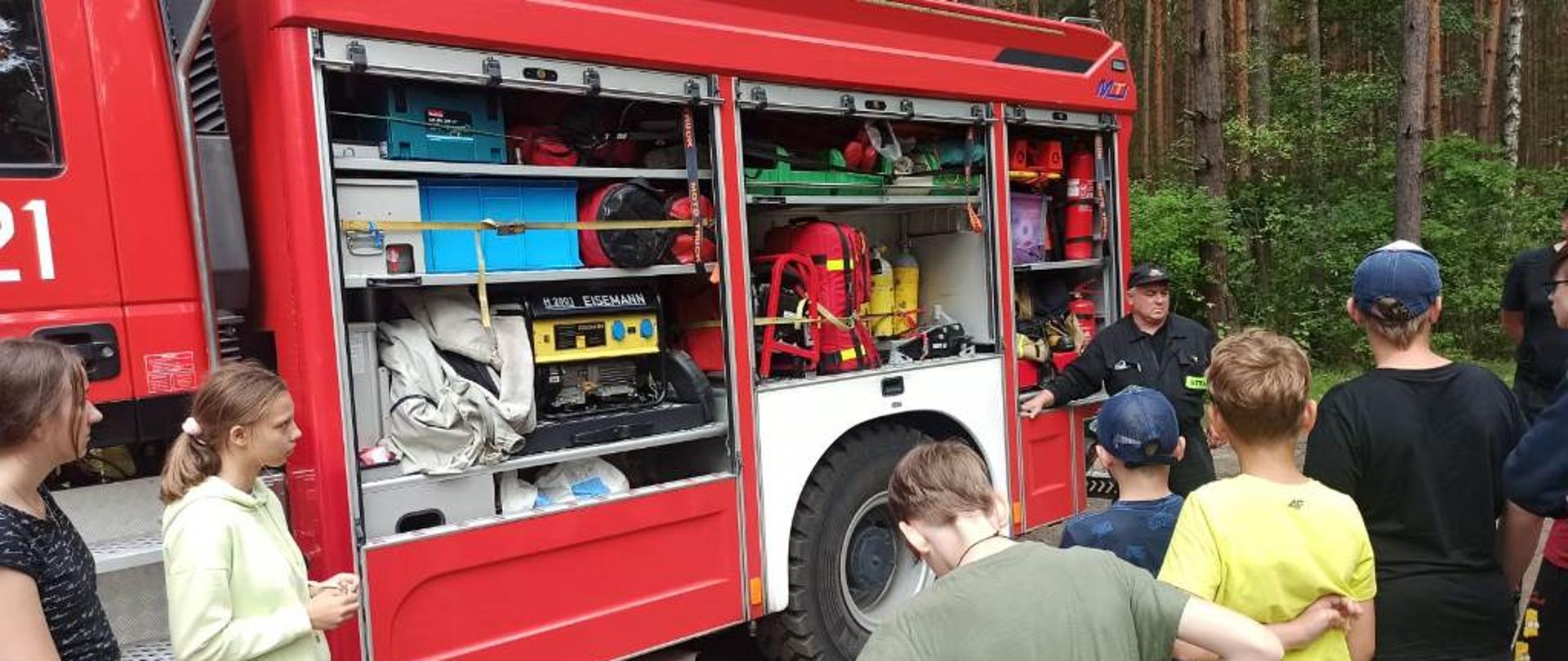 Samochód strażacki o kryptonimie operacyjnym 511-21 z otwartymi skrytkami sprzętowymi, przed samochodem grupa harcerzy słuchająca prelekcji prowadzonej przez strażaków